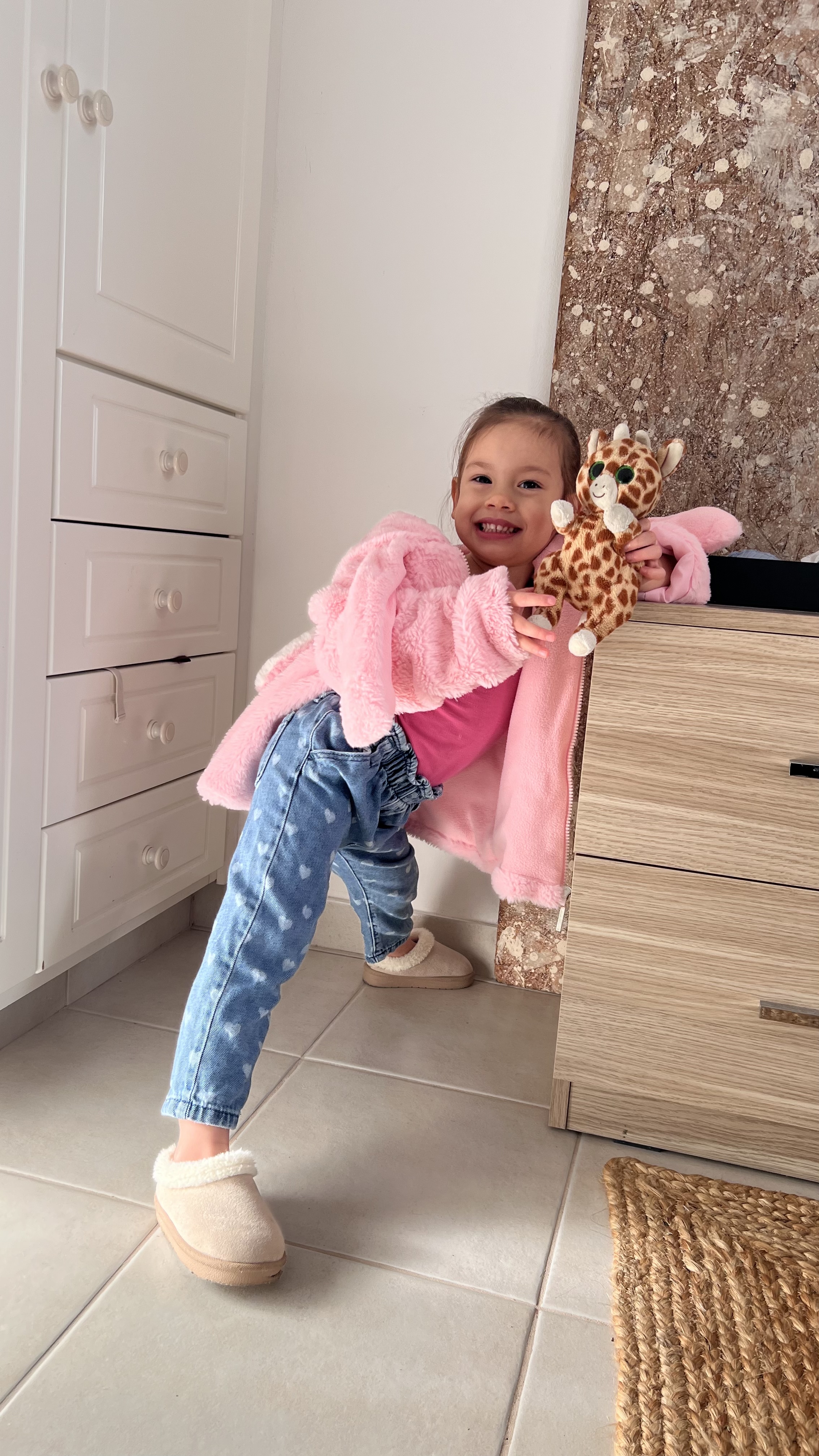 Kiara smiling brightly while holding her toy giraffe, wearing her pink fluffy jacket.